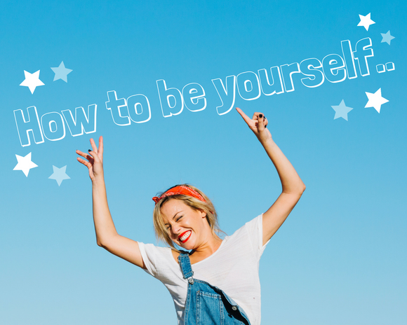 Woman with blonde hair and a red bandana hair tie, wearing a white tshirt and denim dungarees with one strap hanging down is smiling with her hands in the air, pointing to the lettering above her that says 'How to be yourself..' in white