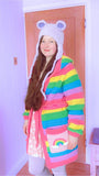 Tamsyn, a white woman with long brown hair is wearing a cute pastel outfit including a VelvetVolcano pastel rainbow striped hooded crochet cardigan that has baby pink pockets with pastel rainbow & cloud motifs, a fluffy lilac teddy bear earflap hat, a baby pink and white cloud print dress and lilac leggings.