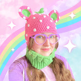 Tamsyn, a white woman with long brown hair is wearing a pastel strawberry theme crochet bear balaclava, lilac heart shaped glasses and purple hoodie.