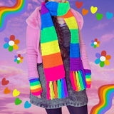 A woman's torso and upper legs are in frame, she is wearing a grey/black acid wash denim pinafore dress, a rainbow striped top with a heart cut out on the chest, a pink ribbed cardigan with a faux fur collar, black tights and a matching neon rainbow striped crochet scarf with rainbow tassels and neon rainbow striped crochet fingerless gloves that are by VelvetVolcano