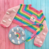 A VelvetVolcano Cropped Crochet Jumper with Pastel Rainbow Body and Baby Pink arms with white cloud appliqué pattern and a pastel rainbow cloud chest pocket and a circular crochet shoulder bag in duck egg blue with pastel rainbow and cloud design and a pastel rainbow striped strap.