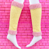 A pair of crochet leg warmers in pastel yellow with baby pink cuffs by VelvetVolcano. The background is pink brick.