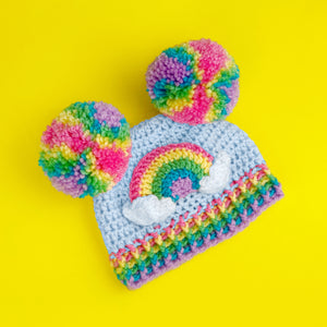 A light blue crochet beanie with pastel rainbow double pom poms, positioned like bear ears, a pastel rainbow and cloud motif on the front of the hat and a pastel striped ribbed brim by VelvetVolcano