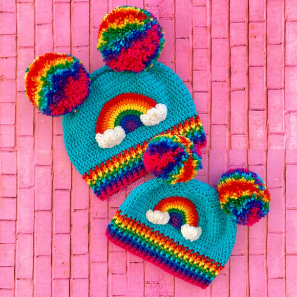 Turquoise crochet beanies with bright rainbow pom poms positioned like ears on the hat, a bright rainbow and white cloud motif on the front of the hat and a rainbow striped rib at the bottom of the hat. - Bright Rainbow Cloud Double Pom Pom Beanie by VelvetVolcano