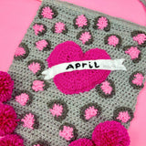Grey crochet pennant wall hanging with bubblegum pink and dark grey leopard print and a hot pink love heart with a white felt banner over the top that says "April" in embroidered black text. The triangular bottom of the wall hanging is trimmed with hot pink pom poms and it hangs from a wooden dowel.