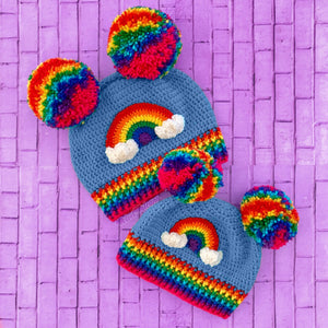 An adult and child size crocheted beanie set, both are dolphin blue with rainbow Pom Poms positioned like bear ears, with rainbow striped ribbed brims and a rainbow and cloud brim on the front of the hat. Hats are by VelvetVolcano.
