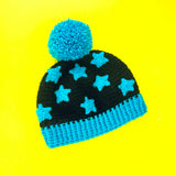 A black crocheted hat with turquoise pom pom, turquoise ribbed brim and a turquoise star applique pattern by VelvetVolcano