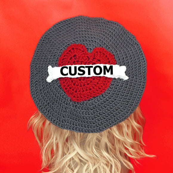 Grey crochet beret with red heart in the centre and a white tattoo style banner over the top that says 