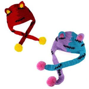 Two crochet cat ear beanies with ear flaps and long ties with pom poms on the ends, designed to look like Frankenstein's Monster cats with a 50/50 style split down the middle with a different colour on each side. The hat on the left is half red, half burgundy with black embroidered stitch details and yellow pom poms and inner ears. The hat on the right is half lilac, half turquoise, with black stitches and bubblegum pink inner ears / pom poms.