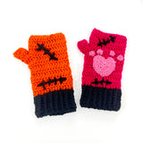 VelvetVolcano FrankenKitty crochet fingerless gloves. One glove is hot pink, the other is orange. Both feature bubblegum pink heart shaped paw prints, black Frankenstein's Monster inspired stitches and black cuffs.