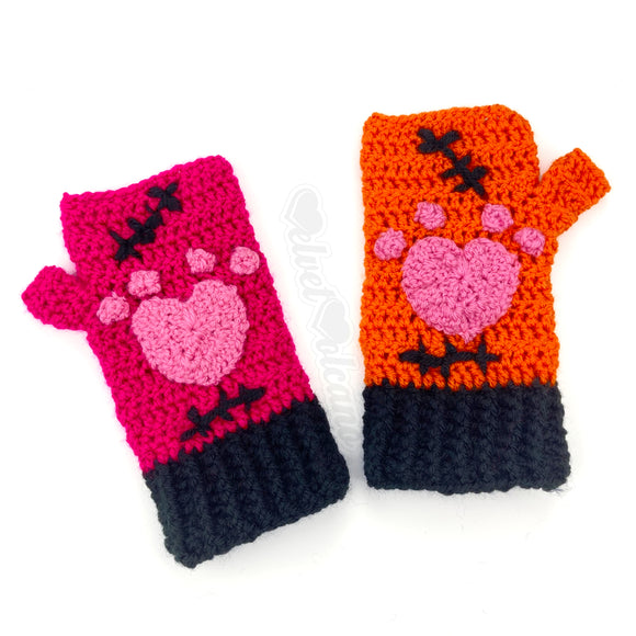 VelvetVolcano FrankenKitty crochet fingerless gloves. One glove is hot pink, the other is orange. Both feature bubblegum pink heart shaped paw prints, black Frankenstein's Monster inspired stitches and black cuffs.