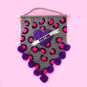 Grey crochet pennant wall hanging with pink and black leopard print and a purple love heart with a white felt banner over the top that says "Custom" in black text. The triangular bottom of the wall hanging is trimmed with purple pom poms and it hangs from a wooden dowel.