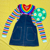 A flat lay outfit photograph on a yellow and white polka dot background. The outfit features a white and rainbow striped jumper, a dark denim pinafore dress a Rainbow striped crocheted Peter Pan Collar and a pastel green crocheted beret with a darker green, white and yellow daisy chain design.
