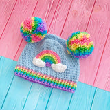 A light blue crochet beanie with pastel rainbow double pom poms, positioned like bear ears, a pastel rainbow and cloud motif on the front of the hat and a pastel striped ribbed brim by VelvetVolcano