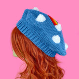 A dolphin blue crocheted beret with a repeating pattern of white clouds and a bright rainbow in the centre with white clouds at both ends by VelvetVolcano