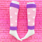 Baby pink crochet leg warmers with white heart pattern and lilac cuffs by VelvetVolcano. Background is pink brick.