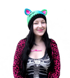 Tamsyn, a white woman with long black hair is wearing a zombie cat inspired "FrankenKitty Beanie", a hot pink and black leopard print cardigan and a black t-shirt with ribcage print