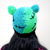 Tamsyn, a white woman with long black hair is wearing a zombie / Frankenstein's Monster inspired crochet cat ear beanie, a pink and black leopard print cardigan and a skeleton tshirt.