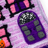 Lilac crochet Haunted House backpack with lavender brick pattern, black window frames with bubblegum pink curtains, a violet scalloped roof, a dark purple door and pumpkin, ghost and spider web accents to the window. The bag also has violet, white, black and dark purple striped straps. Haunted House Backpack by VelvetVolcano