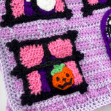 Lilac crochet Haunted House backpack with lavender brick pattern, black window frames with bubblegum pink curtains, a violet scalloped roof, a dark purple door and pumpkin, ghost and spider web accents to the window. The bag also has violet, white, black and dark purple striped straps. Haunted House Backpack by VelvetVolcano