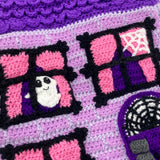 Lilac crochet Haunted House backpack with lavender brick pattern, black window frames with bubblegum pink curtains, a violet scalloped roof, a dark purple door and pumpkin, ghost and spider web accents to the window. The bag also has violet, white, black and dark purple striped straps. Haunted House Backpack by VelvetVolcano