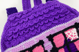 Lilac crochet Haunted House backpack with lavender brick pattern, black window frames with bubblegum pink curtains, a violet scalloped roof, a dark purple door and pumpkin, ghost and spider web accents to the window. The bag also has violet, white, black and dark purple striped straps. Haunted House Backpack by VelvetVolcano