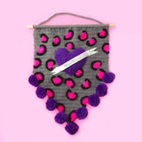 Grey crochet pennant wall hanging with pink and black leopard print and a purple love heart with a white felt banner over the top. The triangular bottom of the wall hanging is trimmed with purple pom poms and it hangs from a wooden dowel.