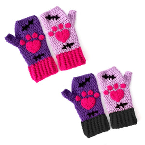 A set of crochet fingerless gloves with a Frankenstein's Monster & Zombie Cat inspired design, with one lilac glove, one violet glove, black embroidered stitch details and hot pink heart shaped paws - NecroKitty Fingerless Gloves by VelvetVolcano