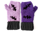 A set of crochet fingerless gloves with a Frankenstein's Monster & Zombie Cat inspired design, with one lilac glove, one violet glove, black embroidered stitch details and cuffs and hot pink heart shaped paws - NecroKitty Fingerless Gloves by VelvetVolcano