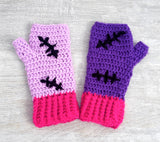 A set of crochet fingerless gloves with a Frankenstein's Monster & Zombie Cat inspired design, with one lilac glove, one violet glove, black embroidered stitch details and hot pink heart shaped paws and cuffs - NecroKitty Fingerless Gloves by VelvetVolcano