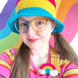 Tamsyn has long light brown hair tied into two low ponytails. She is wearing a crocheted Pansexual Pride bucket hat with 3 thick stripes of Hot Pink, Yellow & Turquoise. She is also wearing rainbow ombre frame glasses; a pink, yellow, black & orange striped tshirt & a yellow dungaree dress. On the strap of the dungaree dress there is a crocheted pansexual rainbow brooch that has white clouds at either end. Behind Tamsyn there is a rainbow coloured thick wavy line painted mural.