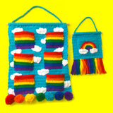 Turquoise crochet wall hanging organiser with white cloud pattern and 6 bright rainbow striped pockets. The wall hanging hangs from a wooden dowel and braided hanger and the bottom is trimmed with 7 pom poms, each in a different rainbow colour. Clouds & Rainbows Hanging Organiser by VelvetVolcano