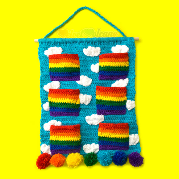 Turquoise crochet wall hanging organiser with white cloud pattern and 6 bright rainbow striped pockets. The wall hanging hangs from a wooden dowel and braided hanger and the bottom is trimmed with 7 pom poms, each in a different rainbow colour. Clouds & Rainbows Hanging Organiser by VelvetVolcano