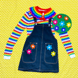 A flat lay outfit photograph on a yellow and white polka dot background. The outfit features a white and rainbow striped jumper, a dark denim pinafore dress a Rainbow striped crocheted Peter Pan Collar and a grass green crocheted beret with rainbow daisy design. The pinafore also has some crocheted rainbow daisy appliques on the pockets.
