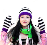 Picture of Tamsyn, a white woman with long black hair wearing a matching black and white striped crochet beanie hat and mittens set with purple cuffs / brim. She is also wearing a neon green crochet scarf with black and white tassels.