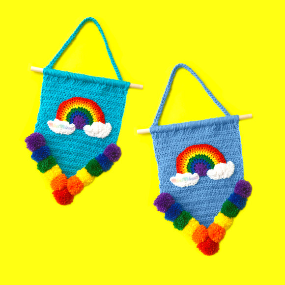 Pennant shape crochet wall hanging in Turquoise or Dolphin Blue with Bright Rainbow and Cloud motif and rainbow pom pom trim, hanging from a wooden dowel and braided hanger. Bright Rainbow Cloud Pennant Wall Hanging by VelvetVolcano