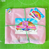 VelvetVolcano Order Packaging showing a baby pink tissue paper wrapped item with a crochet flower tie wrapped around the package and finished with VelvetVolcano rainbow cloud flyer, business cards and pin badge.