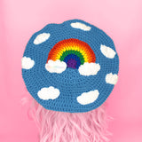 A dolphin blue crocheted beret with a repeating pattern of white clouds and a bright rainbow in the centre with white clouds at both ends by VelvetVolcano