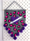 Grey crochet pennant wall hanging with pink and black leopard print and a purple love heart with a white felt banner over the top. The triangular bottom of the wall hanging is trimmed with purple pom poms and it hangs from a wooden dowel.