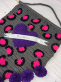 Grey crochet pennant wall hanging with pink and black leopard print and a purple love heart with a white felt banner over the top. The triangular bottom of the wall hanging is trimmed with purple pom poms and it hangs from a wooden dowel.