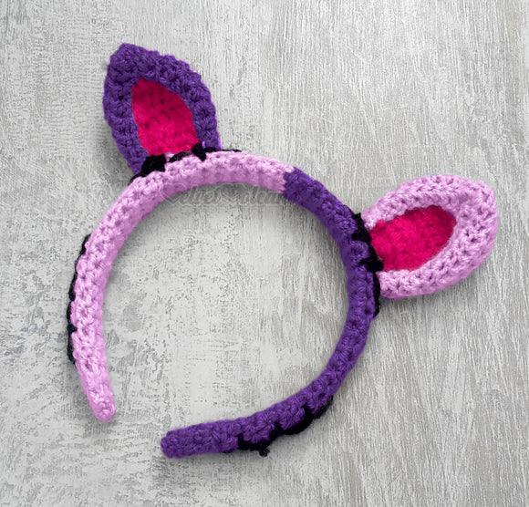 Half lilac and half violet cat ear headband with each ear being the opposite colour to the side it is on. The inner ears are hot pink and there are black embroidered stitch marks to look like the cat has been pieced together. NecroKitty Crochet Headband by VelvetVolcano