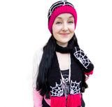 Tamsyn who is a white woman in her 20's with long black hair is wearing a black crochet beanie with light grey spider web design and hot pink ribbed brim, a black scarf with white web detail and hot pink tassels and matching mittens as well as a white to pink ombre cropped biker jacket and a black tshirt with a skeleton print.