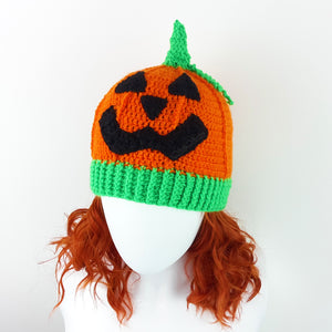 Orange crochet beanie with contrasting neon green ribbed bottom section, neon green stem and a black carved pumpkin face on the main part of the hat. Pumpkin Beanie by VelvetVolcano