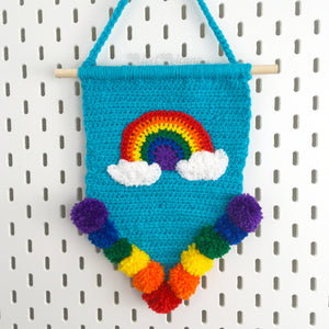 Pennant shape crochet wall hanging in Turquoise or Dolphin Blue with Bright Rainbow and Cloud motif and rainbow pom pom trim, hanging from a wooden dowel and braided hanger. Bright Rainbow Cloud Pennant Wall Hanging by VelvetVolcano