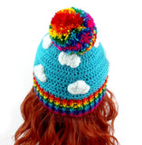 Turquoise crochet beanie with white cloud print, rainbow pom pom and rainbow rib section at the bottom of the hat. Bright Rainbow Cloud Pom Pom Beanie by VelvetVolcano