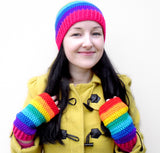 Tamsyn, a white woman with long black hair is wearing a yellow duffle coat, a rainbow striped crochet beanie and rainbow striped crochet fingerless gloves by VelvetVolcano